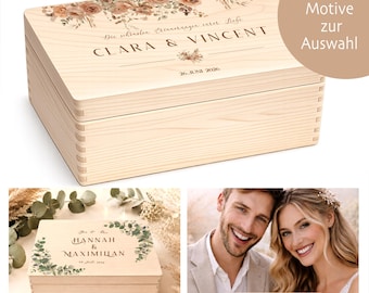 May include: A light-colored wooden box with a hinged lid, featuring floral designs and personalized text. The box is rectangular and has dovetail joints. The text on the box reads "CLARA & VINCENT" and "26. JUNI 2026".