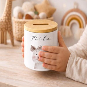 Puede incluir: Hucha de cer&aacute;mica blanca con tapa de madera. La hucha presenta una ilustraci&oacute;n en acuarela de un cerdo y el nombre "Milo" en escritura cursiva. Dise&ntilde;ada para ni&ntilde;os, es un elemento decorativo para una habitaci&oacute;n infantil.