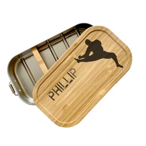 Op de afbeelding: Een lunchbox van roestvrij staal met een bamboe deksel. Het deksel heeft een silhouet van een persoon die een martial arts kick uitvoert en de naam "PHILLIP" erop gegraveerd.