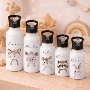 Puede incluir: Cinco botellas de agua blancas con tapas negras, cada una con una ilustración de animal en acuarela y un nombre: Ezra, Milo, Isla, Oliver y Nora. Los animales incluyen un zorro, un cerdo, un oso, un mapache y un conejo. Las botellas son para niños.