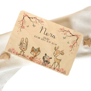 May include: Wooden birth announcement plaque with a pink and white floral design. The plaque features a bunny, fox, raccoon, and deer with floral headbands. The text on the plaque reads "Nora 25.04.2023 18:37 Uhr 4220 g 56 cm 38,5 cm".