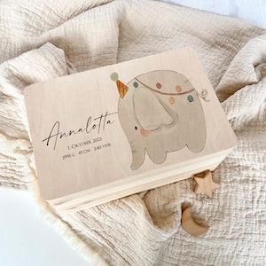 May include: A wooden keepsake box with a light wood finish. The box features a hand-painted illustration of a grey elephant with a party hat and colourful decorations. The box is personalised with the name "Annalotta" and the text "7. Oktober 2023, 2998 g, 49 cm, 2:40 Uhr".