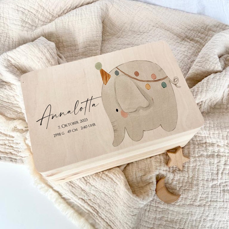 May include: A wooden keepsake box with a light wood finish. The box features a hand-painted illustration of a grey elephant with a party hat and colourful decorations. The box is personalised with the name "Annalotta" and the text "7. Oktober 2023, 2998 g, 49 cm, 2:40 Uhr".