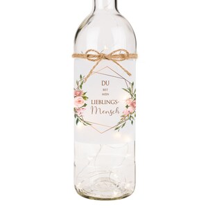 May include: A clear glass bottle with a floral design and the text "DU BIST MEIN LIEBLINGS-MENSCH" in a gold geometric frame. The bottle is decorated with a brown twine bow and has a string of white lights inside.