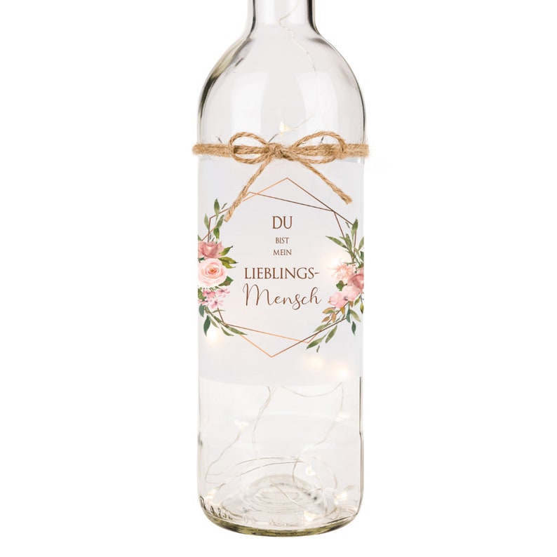 May include: A clear glass bottle with a floral design and the text "DU BIST MEIN LIEBLINGS-MENSCH" in a gold geometric frame. The bottle is decorated with a brown twine bow and has a string of white lights inside.