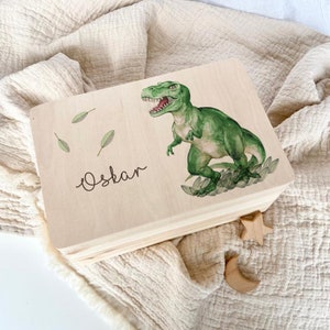 Może przedstawiać: Jasna drewniana skrzynka z pokrywą na zawiasach, ozdobiona ilustracją zielonego Tyrannosaurus Rexa i imieniem "Oskar" w ozdobnym kroju pisma. Skrzynka jest ozdobiona zielonymi liśćmi i leży na beżowej tkaninie.