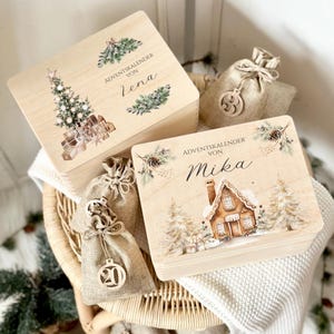 Può includere: Scatole di calendario dell'Avvento in legno con illustrazioni natalizie. Una scatola presenta un design ad albero di Natale, l'altra una casa di pan di zenzero. Sono presenti anche sacchetti di iuta con ornamenti in legno.
