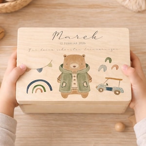 May include: A light brown wooden keepsake box featuring a bear illustration in a green jacket, a rainbow, and a car. The lid is printed with the name "Marek" and the date "12. Februar 2026". The box is held by two hands.