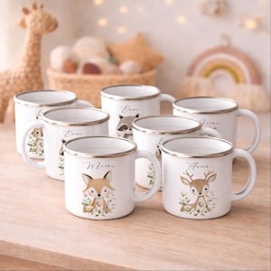 Op de afbeelding: Een verzameling witte emaille mokken met zilverkleurige randen, elk voorzien van een unieke aquarelillustratie van een bosdier zoals een vos, hert of wasbeer, omringd door bloemaccenten en gepersonaliseerd met een naam in een sierlijk handschrift.