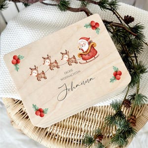 Könnte beinhalten: Eine Holzkiste mit Weihnachtsmotiv, auf der der Weihnachtsmann in einem roten Schlitten von Rentieren gezogen wird. Die Kiste hat einen hellen Holzgrund mit roten Beeren und grünen Blättern. Der Text "Frohe Weihnachten Johanna" ist auf die Kiste gedruckt.