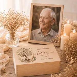 Puede incluir: Una caja rectangular de madera clara con un diseño de árbol grabado, el texto "GELIEBT & UNVERGESSEN", el nombre "Lorenz", un pequeño icono de corazón y las fechas "14. April 1957 - 13. Mai 2026" en la tapa.