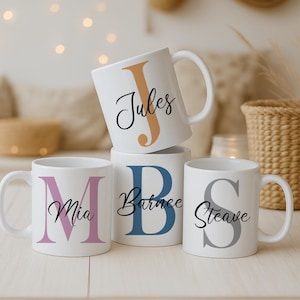 Peut inclure: Quatre mugs en céramique blanche personnalisés avec des noms et des initiales. Les tasses présentent les noms "Jules", "Mia", "Barnee" et "Steave" en écriture cursive. Les initiales sont de différentes couleurs : orange, rose, bleu et gris.