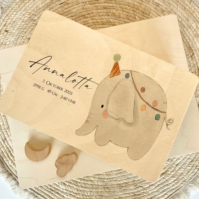 May include: A wooden plaque with a hand-painted illustration of a grey elephant wearing a party hat. The plaque is decorated with colourful dots and has the text "Annalotta 7. OKTOBER 2023 2998 G 49 CM 2:40 UHR" written on it.