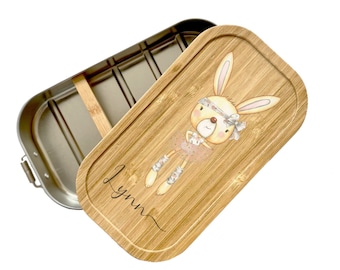 Peut inclure: Une boîte à lunch en acier inoxydable avec un couvercle en bambou. Le couvercle présente une illustration de dessin animé d'un lapin ballerine avec un tutu rose et le nom "Lynn" écrit en cursive.