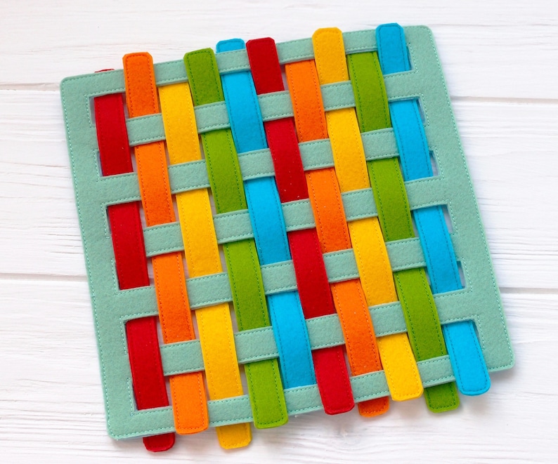 May include: A woven felt mat with colorful strips in red, orange, yellow, green, blue, and turquoise. The strips are woven together in a criss-cross pattern.