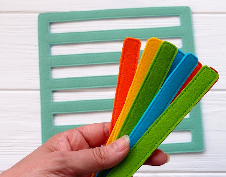 May include: A set of six felt strips in various colors, including green, blue, orange, and yellow. The strips are arranged in a fan shape and held in a hand.
