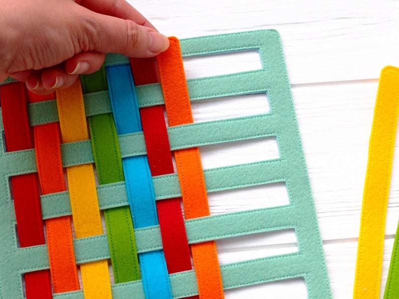 May include: A woven felt mat with a rainbow pattern. The mat is made of strips of felt in red, orange, yellow, green, blue, and turquoise. The strips are woven together to create a checkered pattern. The mat is being held by a hand.
