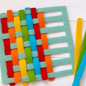 May include: A green felt board with cutouts for weaving colorful felt strips. The strips are red, orange, yellow, green, blue, and purple.