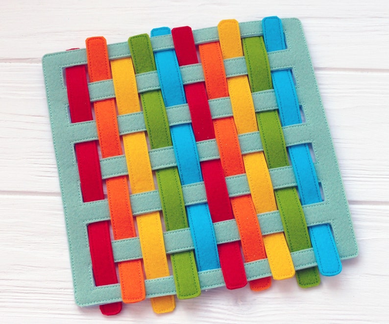 May include: A woven felt mat with colorful strips in red, orange, yellow, green, and blue. The strips are woven together in a criss-cross pattern on a light blue background.