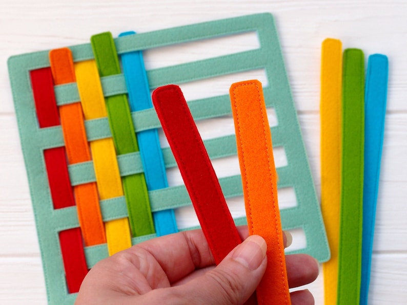May include: A felt weaving activity set with a blue base and colorful felt strips. The strips are red, orange, yellow, green, and blue. The set is designed for children to practice weaving skills.