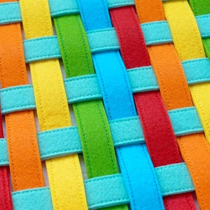 May include: A woven fabric mat with colorful stripes in red, orange, yellow, green, blue, and turquoise. The stripes are woven together to create a checkered pattern.