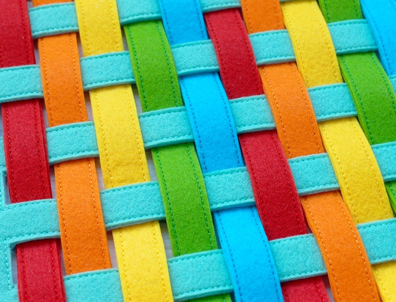May include: A woven fabric mat with colorful stripes in red, orange, yellow, green, blue, and turquoise. The stripes are woven together to create a checkered pattern.