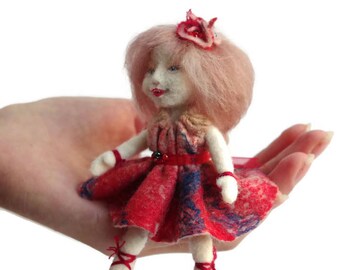 May include: A small felt doll with pink hair, wearing a red and blue dress with a red sash and a flower crown.