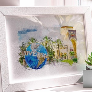 May include: A white framed watercolor painting of a globe with the word "Universal" on it, surrounded by palm trees and a building with an archway. The painting is on a white shelf with a small potted succulent and a business card that says "Happy Place" in front of it.