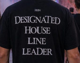 May include: Black t-shirt with the text "DESIGNATED HOUSE LINE LEADER" in white. The image shows a person walking in a crowd at an outdoor event. The background includes other people, buildings, and street lights.