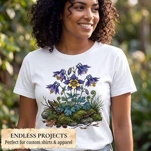 May include: A white t-shirt featuring a detailed graphic illustration of purple and yellow columbine flowers growing from a mossy rock base with green foliage, small pinecones, and fallen oak leaves centered on the front of the garment.