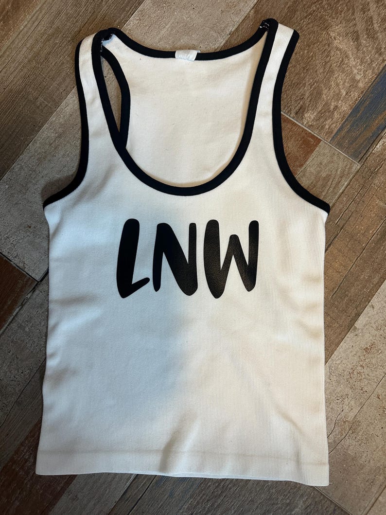 May include: White ribbed tank top with black trim and the letters "LNW" printed in black across the front. The tank top has a racerback design.