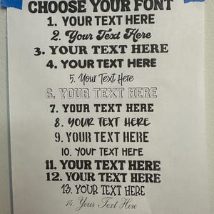 May include: A white paper sheet with the black text CHOOSE YOUR FONT at the top, followed by a numbered list from 1 to 14, each displaying the phrase YOUR TEXT HERE in a different black typeface style, secured to a wall with blue tape.