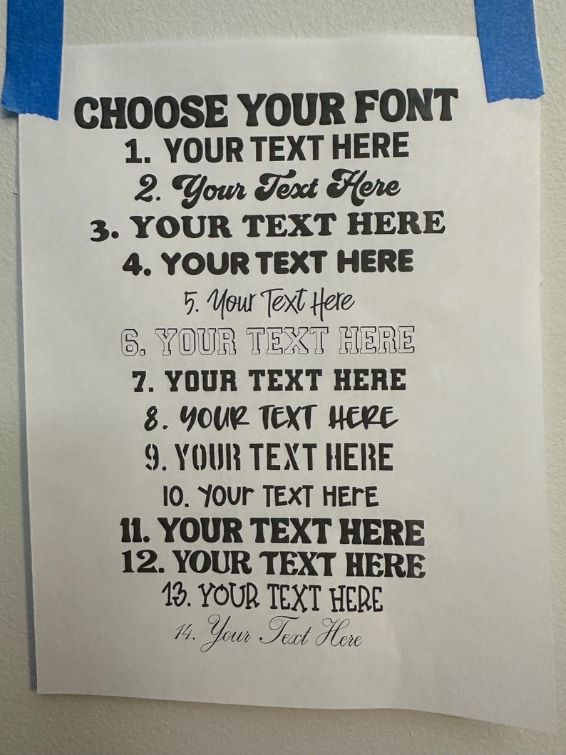May include: A white paper sheet with the black text CHOOSE YOUR FONT at the top, followed by a numbered list from 1 to 14, each displaying the phrase YOUR TEXT HERE in a different black typeface style, secured to a wall with blue tape.