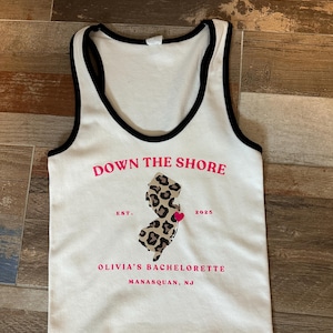 May include: White tank top with black trim and the text "DOWN THE SHORE" in pink. The shirt features a leopard print outline of New Jersey with a pink heart, the text "OLIVIA'S BACHELORETTE" and "MANASQUAN, NJ".