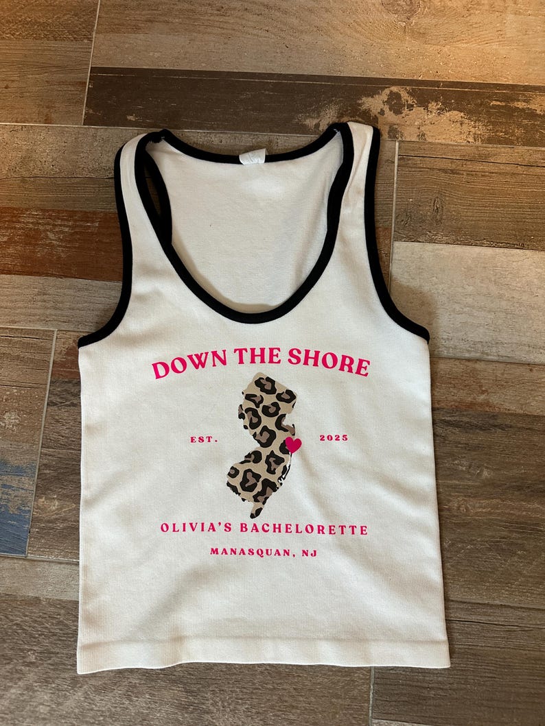 May include: White tank top with black trim and the text "DOWN THE SHORE" in pink. The shirt features a leopard print outline of New Jersey with a pink heart, the text "OLIVIA'S BACHELORETTE" and "MANASQUAN, NJ".
