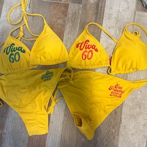 May include: Two yellow string bikinis featuring triangle tops and side-tie bottoms. One set displays green text reading Viva 60 and San Pancho 049. The other set features red text reading Viva 60 and San Pancho 2025. Both sets have thin straps.