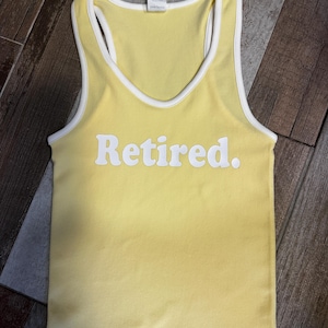 May include: Yellow tank top with white trim and the word "Retired" in white block letters. The top has a racerback design and is made of a soft, ribbed material. The top is a light yellow color.