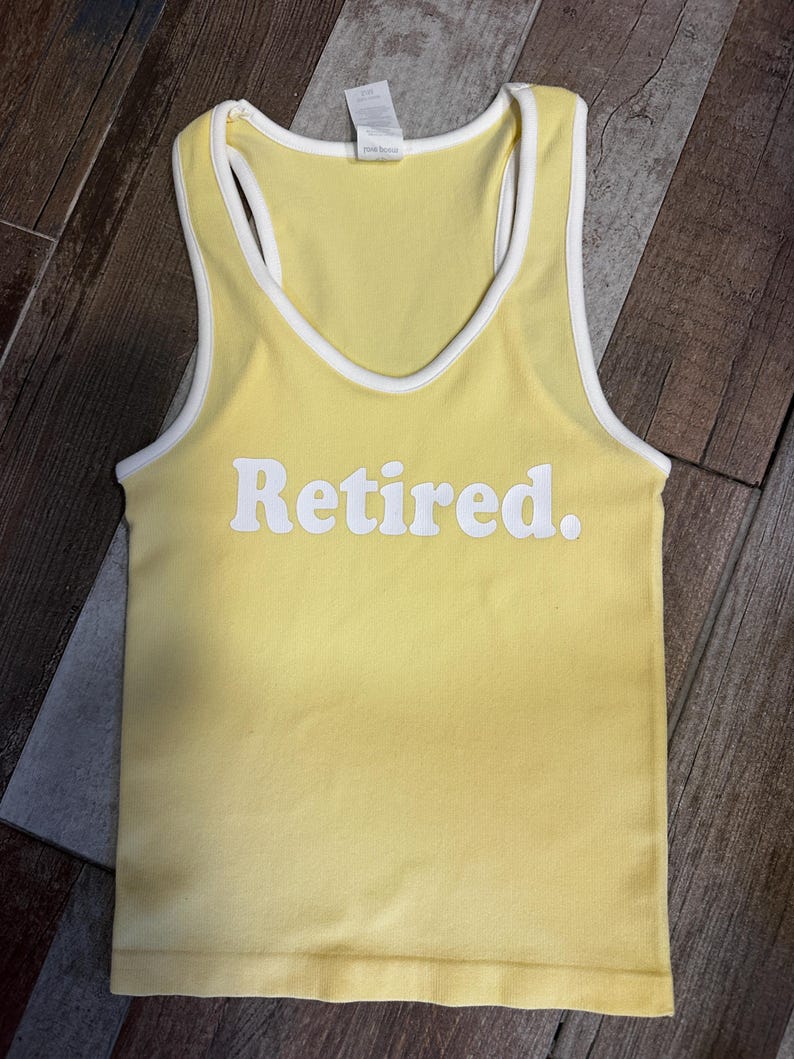 May include: Yellow tank top with white trim and the word "Retired" in white block letters. The top has a racerback design and is made of a soft, ribbed material. The top is a light yellow color.
