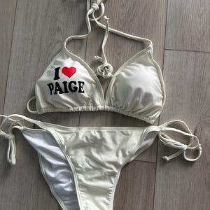 May include: A white two-piece bikini set featuring a triangle top with thin halter neck ties and a matching bottom with side tie closures. The left cup of the top displays the text I followed by a red heart symbol and the name PAIGE in black.