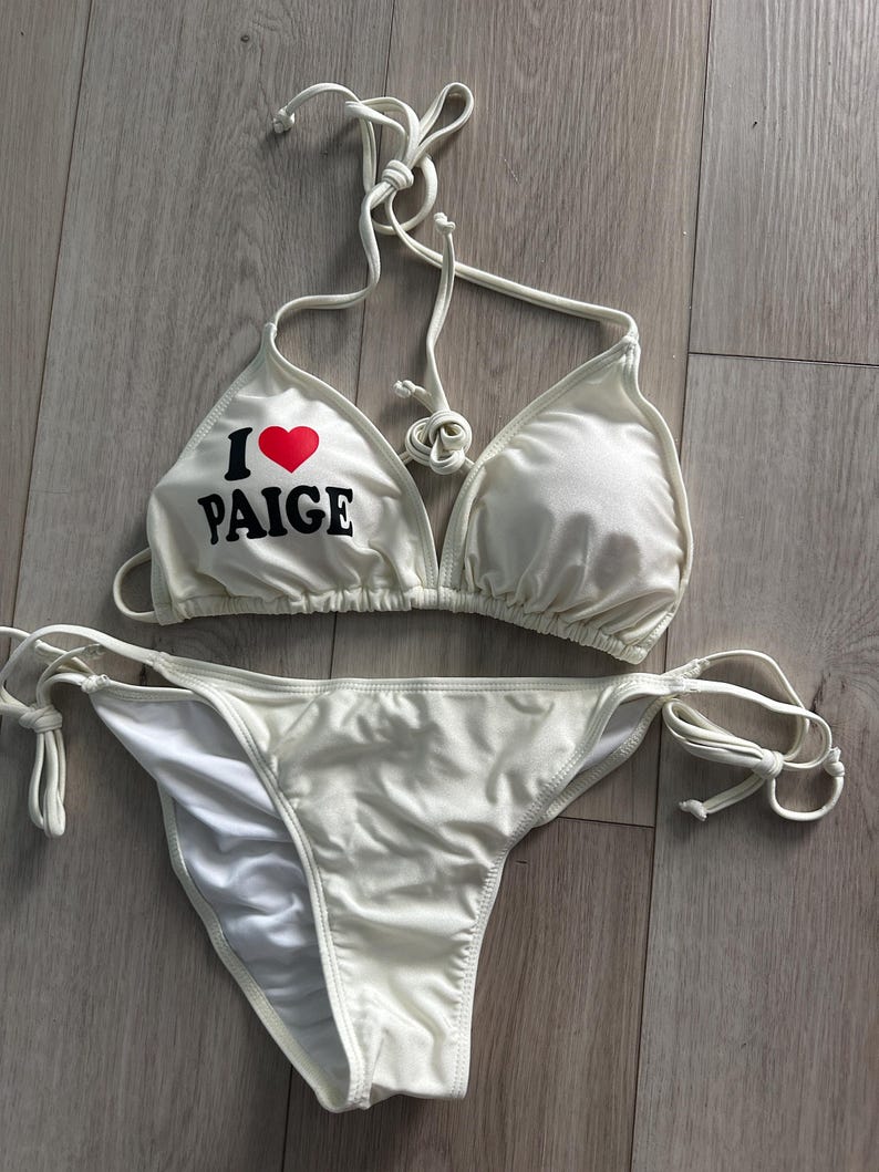May include: A white two-piece bikini set featuring a triangle top with thin halter neck ties and a matching bottom with side tie closures. The left cup of the top displays the text I followed by a red heart symbol and the name PAIGE in black.
