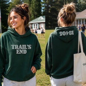 May include: A dark green hoodie with the text "TRAIL'S END" in white block letters on the front. The back of the hoodie has the number "18405" in white. A cream-colored tote bag with "TRAIL'S END CAMP" is also visible.