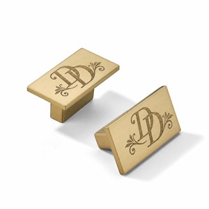 May include: Two rectangular cabinet knobs with a brushed gold finish. Each knob features a centred, engraved monogram of the letters DD, accented with decorative flourishes on the left and right sides of the lettering, set on a flat surface.