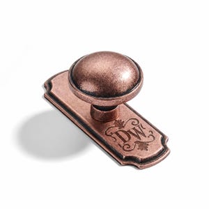 May include: A copper-toned metal cabinet knob featuring a round, domed head attached to a rectangular backplate with rounded, scalloped edges. The backplate is engraved with the letters DW surrounded by decorative flourishes and scrollwork.