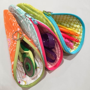 May include: Three colorful zippered pouches with different patterns and designs. The pouches are open and show various items inside, including pens, pencils, a purple tool, and a green rotary cutter.