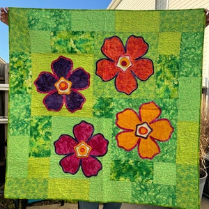 May include: A green quilt with four large, colorful flowers. The flowers are in shades of red, orange, purple, and pink. The quilt is made of patchwork fabric and has a quilted design.