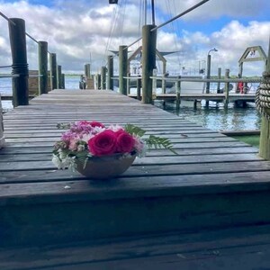 Pu&ograve; includere: Un molo di legno con un mazzo di fiori rosa e bianchi in un vaso marrone. Il molo conduce a una barca nell'acqua.