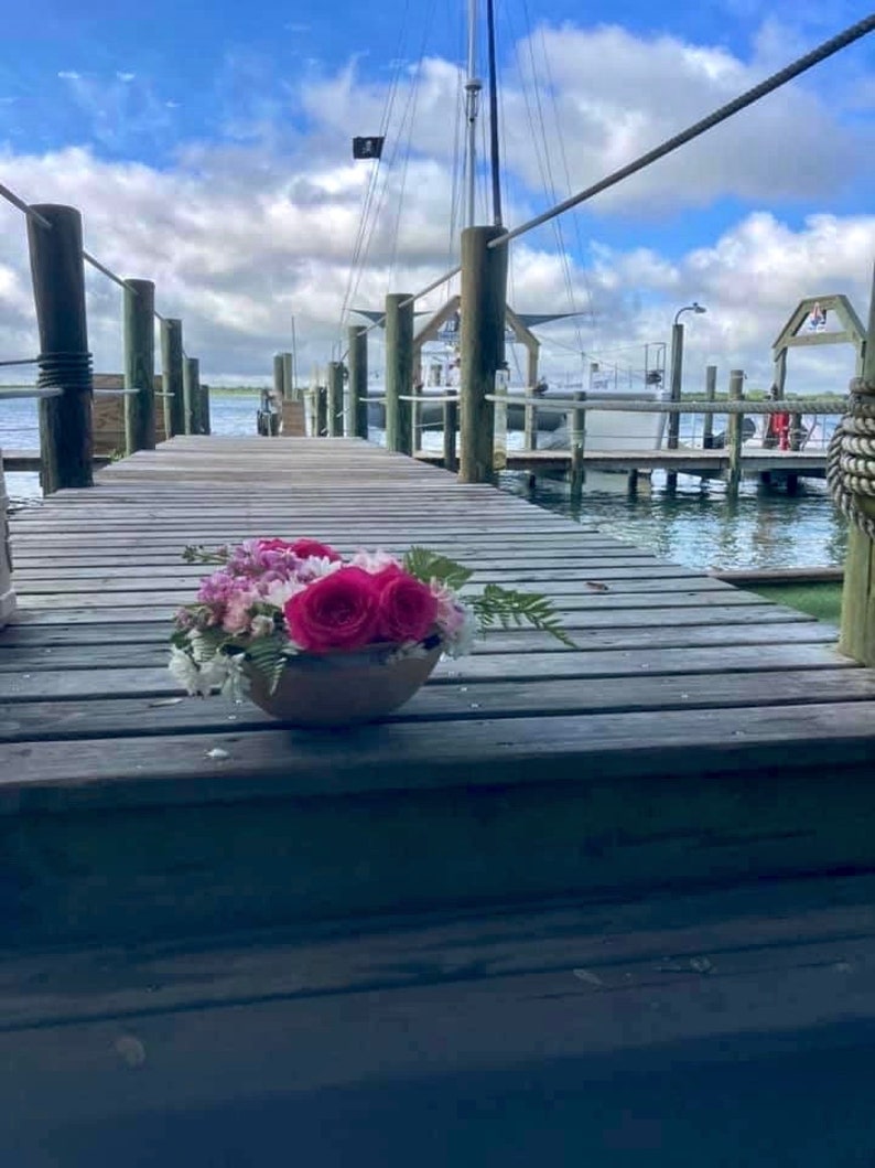 Pu&ograve; includere: Un molo di legno con un mazzo di fiori rosa e bianchi in un vaso marrone. Il molo conduce a una barca nell'acqua.