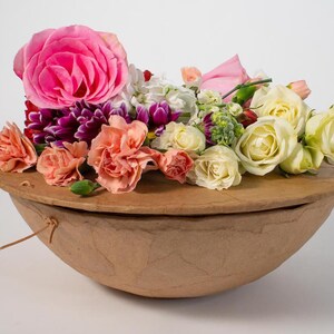 Pu&ograve; includere: Una ciotola di carta marrone con un coperchio, piena di una variet&agrave; di fiori, tra cui rose rosa, garofani bianchi e garofani color pesca.
