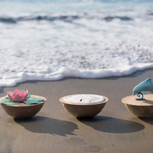 Pode incluir: Tr&ecirc;s tigelas decorativas em uma praia de areia com ondas do oceano ao fundo. As tigelas s&atilde;o feitas de papel mach&ecirc; marrom e cada uma tem um design diferente na parte superior. A primeira tigela tem uma flor de papel rosa e verde, a segunda tigela tem uma concha de mar de papel branco e a terceira tigela tem um golfinho de papel azul.