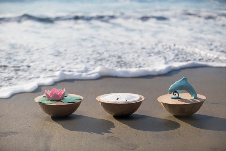 Pode incluir: Tr&ecirc;s tigelas decorativas em uma praia de areia com ondas do oceano ao fundo. As tigelas s&atilde;o feitas de papel mach&ecirc; marrom e cada uma tem um design diferente na parte superior. A primeira tigela tem uma flor de papel rosa e verde, a segunda tigela tem uma concha de mar de papel branco e a terceira tigela tem um golfinho de papel azul.